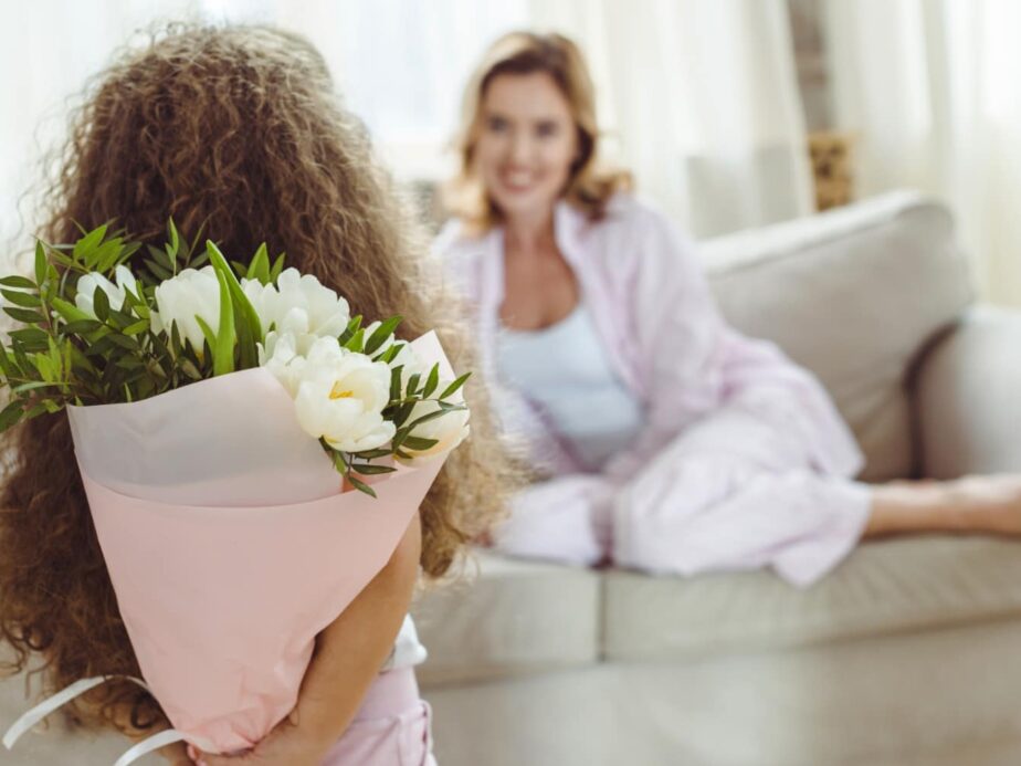 girl with flowers behind her back standing in foreground, white mom on couch in background
