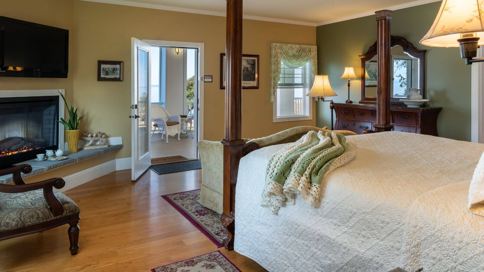 maple four-poster queen bed facing private enclosed patio with wicker furniture, fireplace, flat-screened tv on wall, maple dresser with mirror