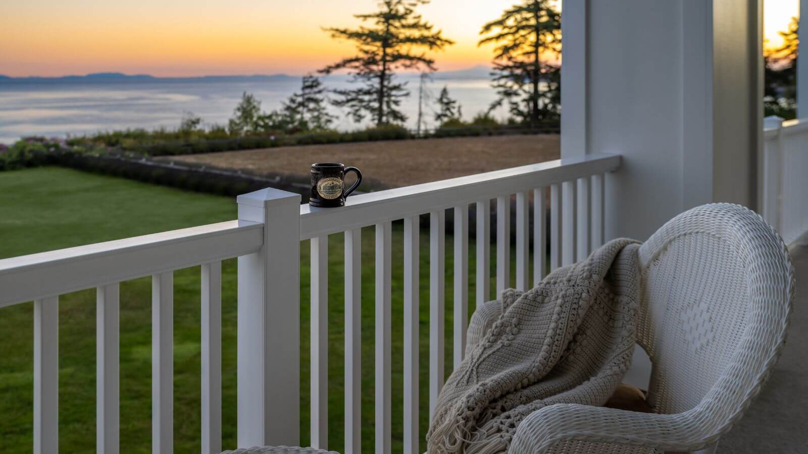 porch facing ocean at sunset, coffee mug sitting on white railing, white wicker chair with beige blanket,