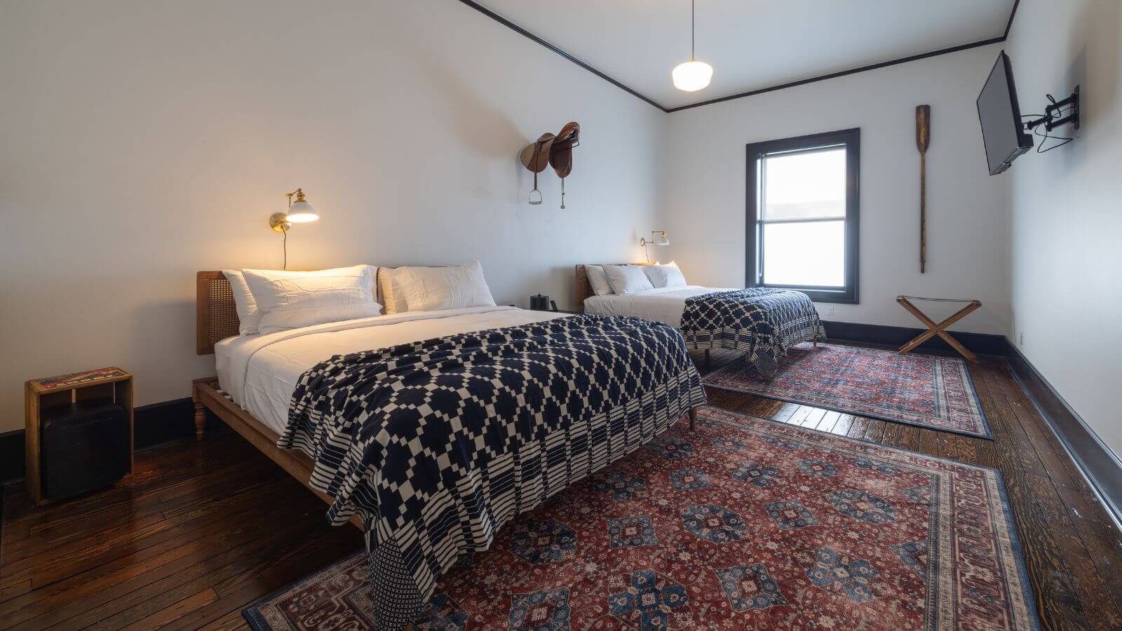 narrow room with king and queen bed with black and white comforters, white walls, hardwood floor and throw rugs, flat-screen television hanging, saddle on wall