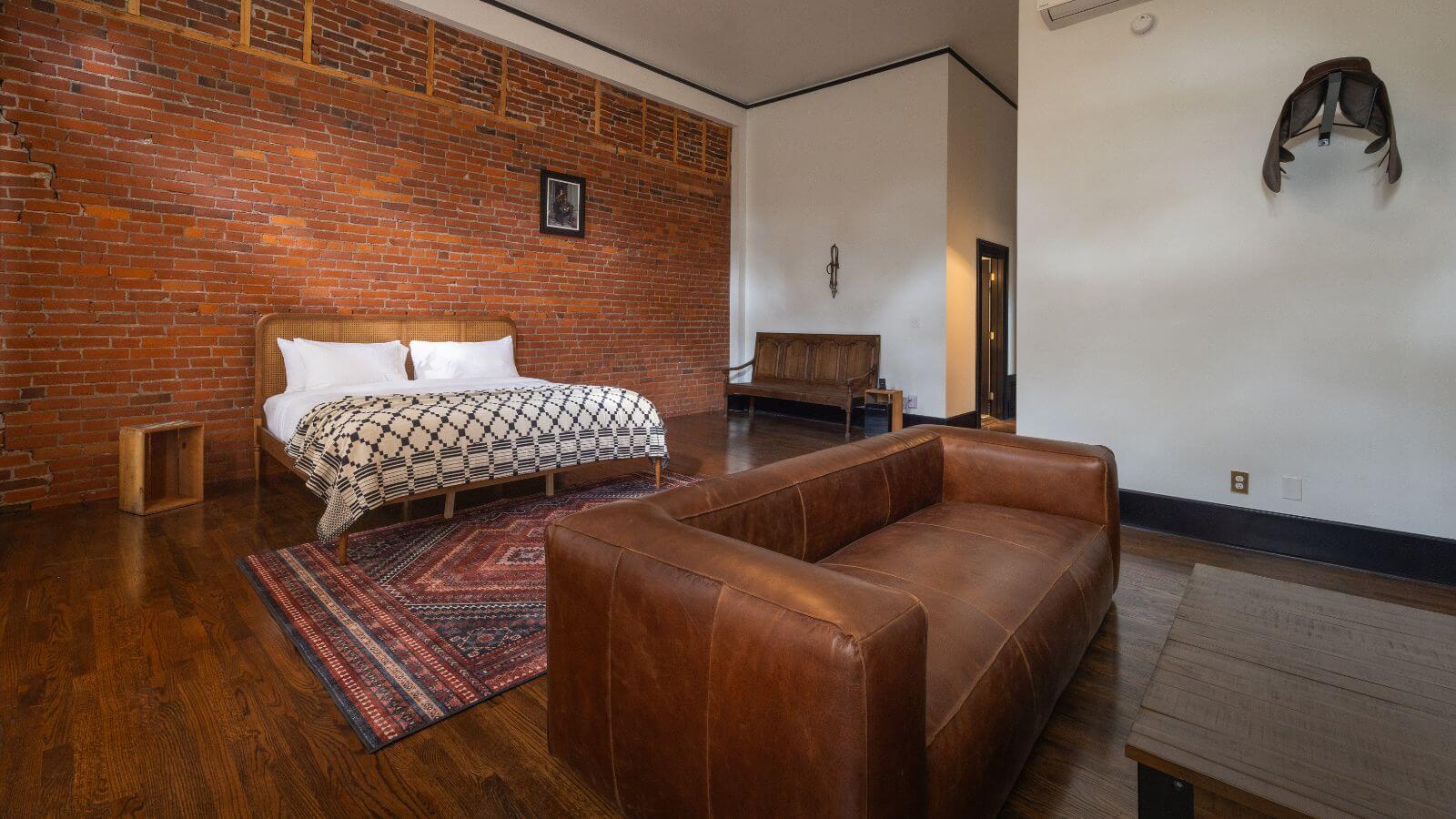 large room with hardwood floor and throw rug, red brick wall and white painted walls, king bed with black and cream comforter, leather couch with wood coffee table