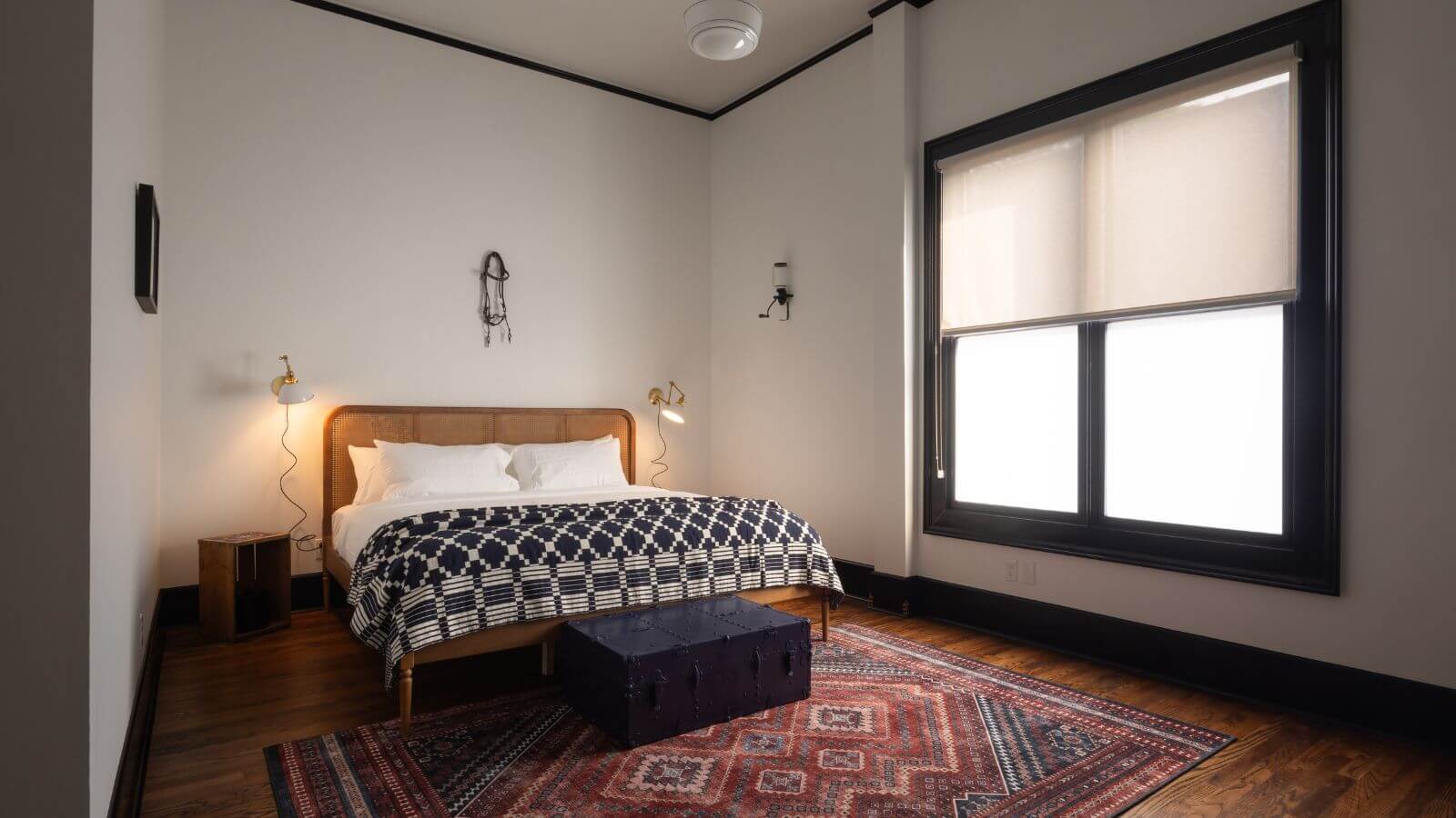 white room with hardwood floors and throw rug, queen bed with black and white comforter with bench at the end, lights hanging on walls next to bed