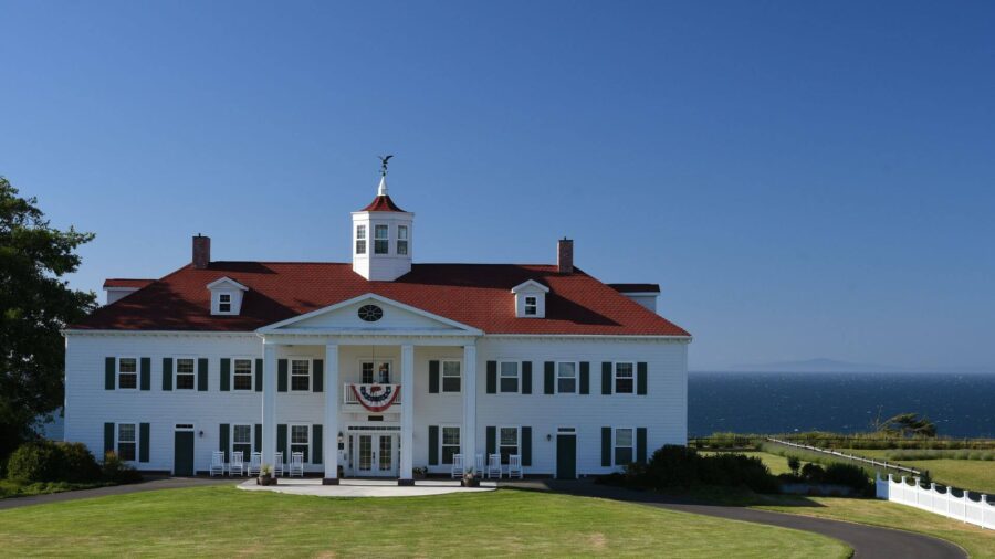 white two-story building with red roof, many windows with black shutters, porch with white rocking chairs