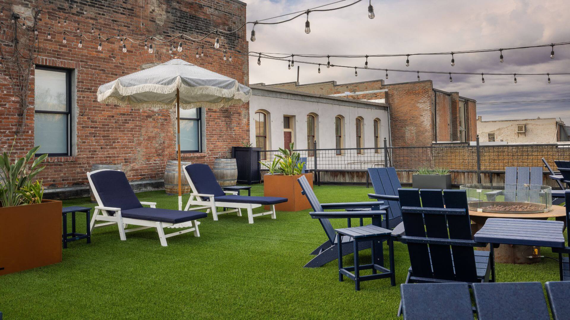 red brick build rooftop set-up with artificial grass with white lounge chairs with blue cushions, lights strung overtop, firepit with blue adirondack chairs