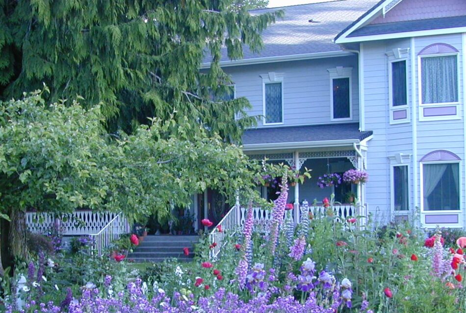Exterior view of two-story property painted light lavender with white trim and surrounded by large green trees and colorful flower gardens